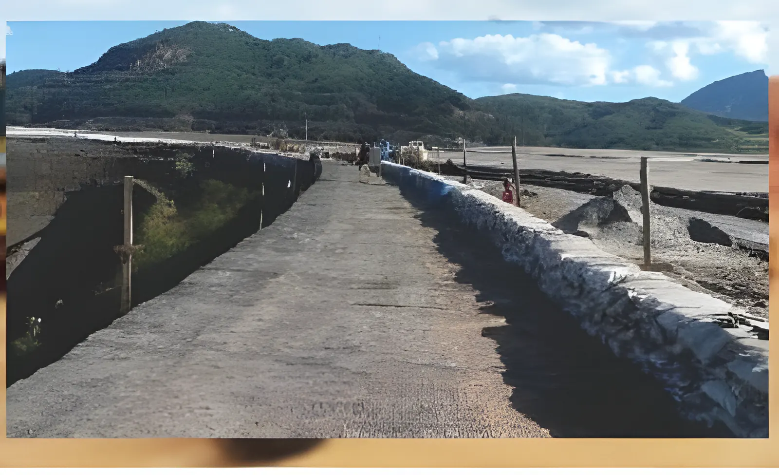 Pista sobre a crista de barragem, com enrocamento na lateral e montanhas ao fundo