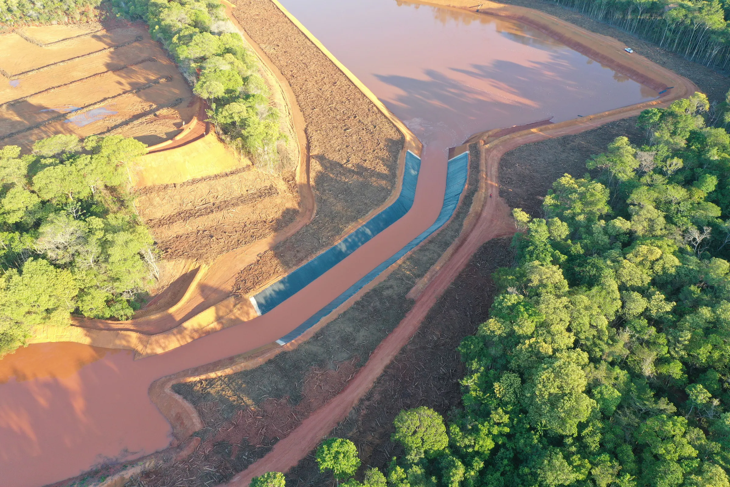 Vista aérea de reservatório de água avermelhada cercado por floresta, com canal revestido e taludes em bancadas